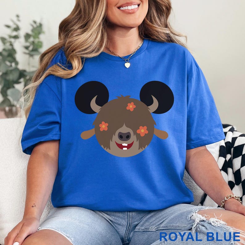 May include: Royal blue t-shirt featuring a cartoon character with large black ears, brown fur, and orange flower accents. The character has a smiling face with two white teeth. The text "ROYAL BLUE" is at the bottom of the image.