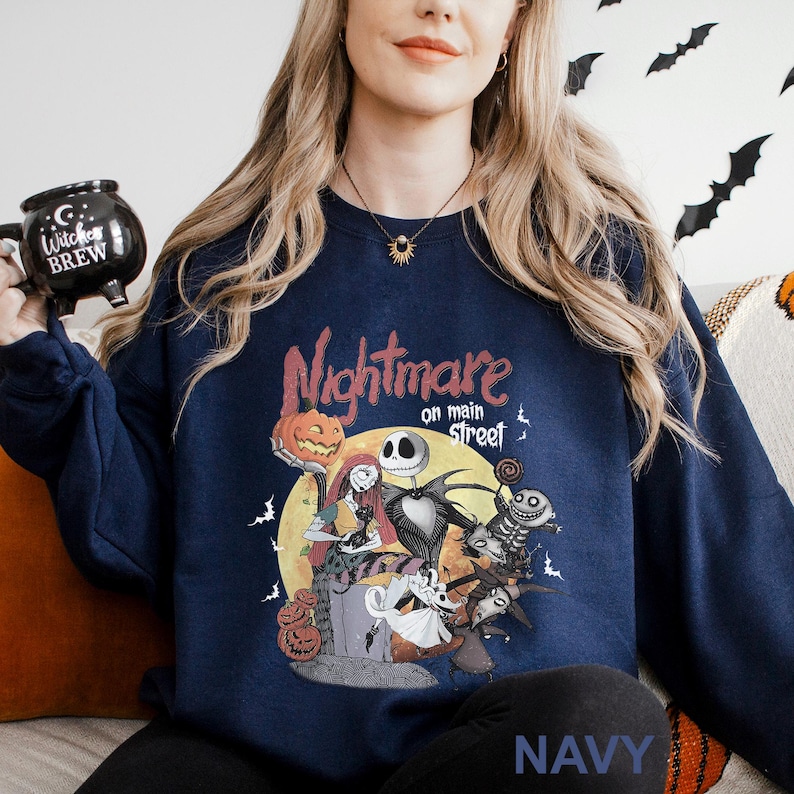 Może przedstawiać: Granatowa bluza z nadrukiem z filmu "Miasteczko Halloween". Projekt zawiera Jacka Skellingtona, Sally i inne postacie na tle pełni księżyca. Tekst "Nightmare on main street" znajduje się nad postaciami. Słowo "NAVY" znajduje się na dole.