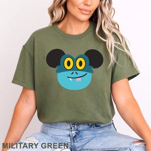 May include: Military green t-shirt featuring a cartoon monster face with black ears, yellow eyes, and a blue face. The monster has a small pink tongue and a smiling mouth. The text "MILITARY GREEN" is at the bottom.