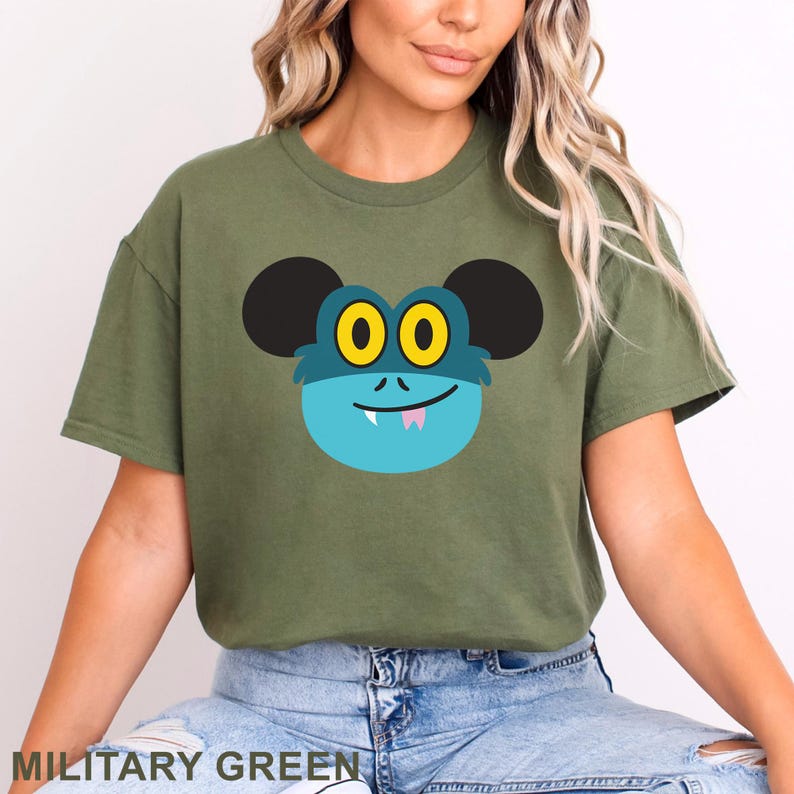 May include: Military green t-shirt featuring a cartoon monster face with black ears, yellow eyes, and a blue face. The monster has a small pink tongue and a smiling mouth. The text "MILITARY GREEN" is at the bottom.