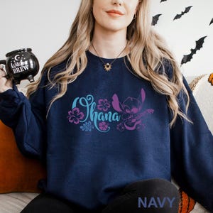 May include: Navy blue sweatshirt featuring the word "Ohana" in teal and purple, with a floral design and a cartoon character playing a ukulele. The word "NAVY" is printed in white at the bottom of the sweatshirt.