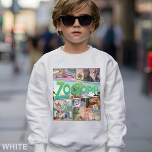 May include: A white sweatshirt featuring a colorful comic strip design with characters from the animated movie "Zootopia." The word "Zootopia" is prominently displayed in green and teal. The sweatshirt is worn by a person wearing sunglasses.