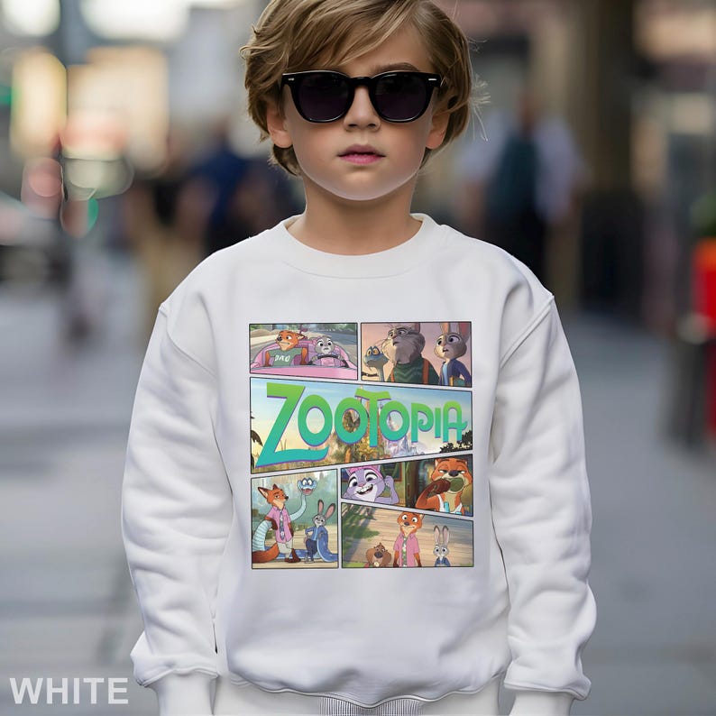 May include: A white sweatshirt featuring a colorful comic strip design with characters from the animated movie "Zootopia." The word "Zootopia" is prominently displayed in green and teal. The sweatshirt is worn by a person wearing sunglasses.