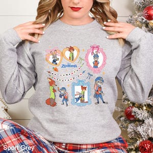 以下が含まれることがあります： ディズニー映画「ズートピア」のキャラクターが描かれたスポーツグレーのスウェットシャツ。ニック・ワイルド、ジュディ・ホップス、その他のキャラクターがフレームやハート型のメガネに入ったデザインです。「Welcome to Zootopia」の文字も含まれています。