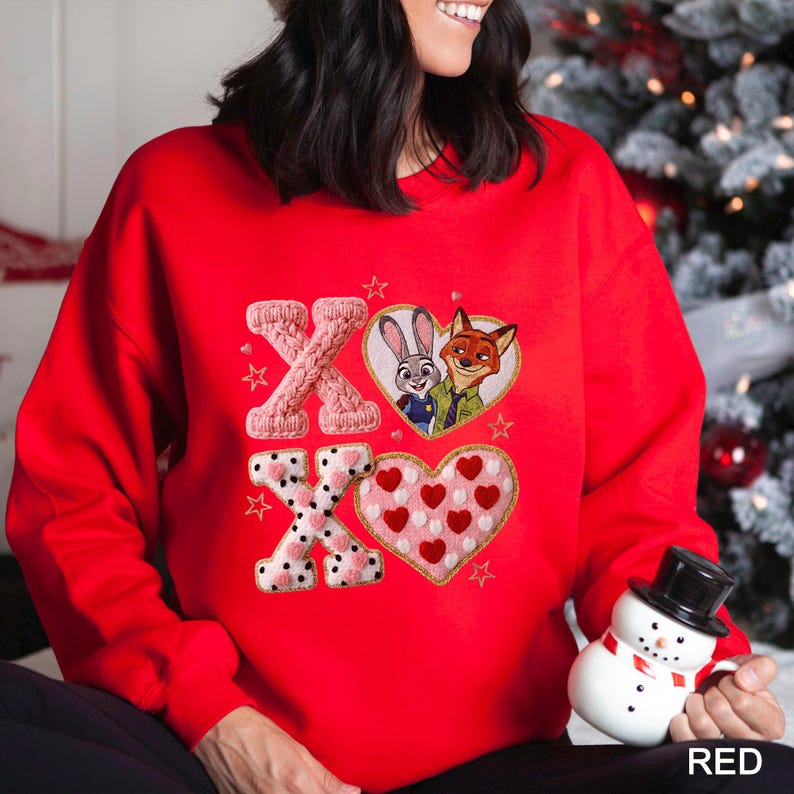 May include: A red sweatshirt with an embroidered design featuring the characters Judy Hopps and Nick Wilde from Zootopia inside a heart. The design also includes the letters "X" and "O" with heart and star accents.