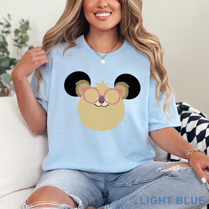 May include: Light blue t-shirt featuring a cartoon koala face wearing pink glasses. The koala has black ears and a tan face. The words "LIGHT BLUE" are printed at the bottom of the shirt.