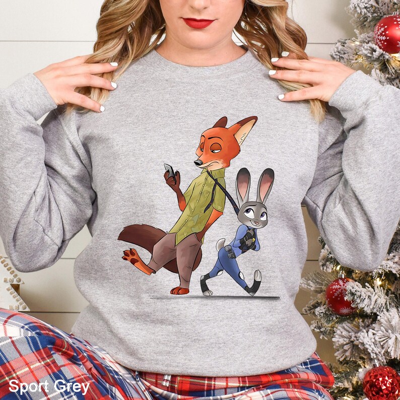 May include: Sport Grey sweatshirt featuring a cartoon illustration of Nick Wilde the fox and Judy Hopps the rabbit from Zootopia. Nick is holding a phone, and Judy is pulling him along. The sweatshirt is a light gray color.