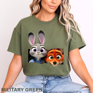 May include: Military green t-shirt featuring a graphic of Judy Hopps and Nick Wilde from Zootopia. The cartoon characters are in a rectangular cutout. The text "MILITARY GREEN" is at the bottom of the shirt.