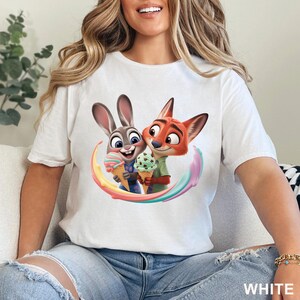 May include: White t-shirt featuring a colorful graphic of Judy Hopps and Nick Wilde from Zootopia, each holding an ice cream cone. The design includes a rainbow-colored swirl. The word "WHITE" is printed in the lower right corner.