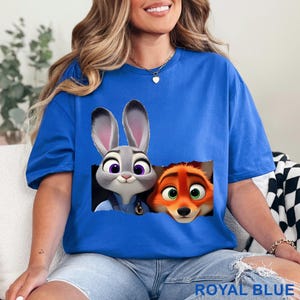 May include: Royal blue t-shirt featuring a graphic of a gray bunny and an orange fox. The bunny has purple eyes and the fox has green eyes. The text "ROYAL BLUE" is at the bottom of the shirt.