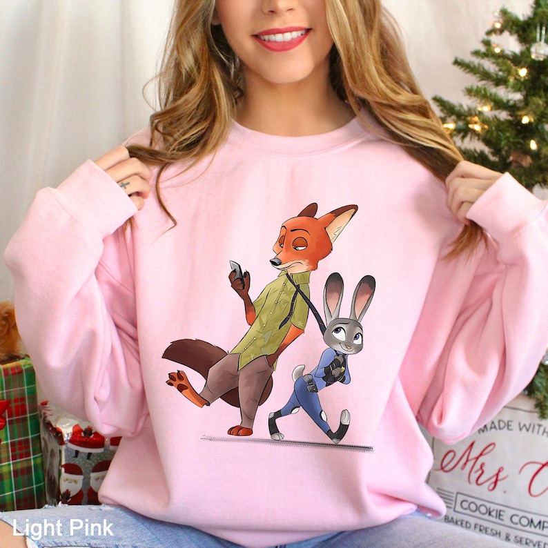 May include: Light pink sweatshirt featuring a cartoon illustration of a fox and a rabbit. The fox is holding a phone and the rabbit is walking. The text "Light Pink" is at the bottom of the image.
