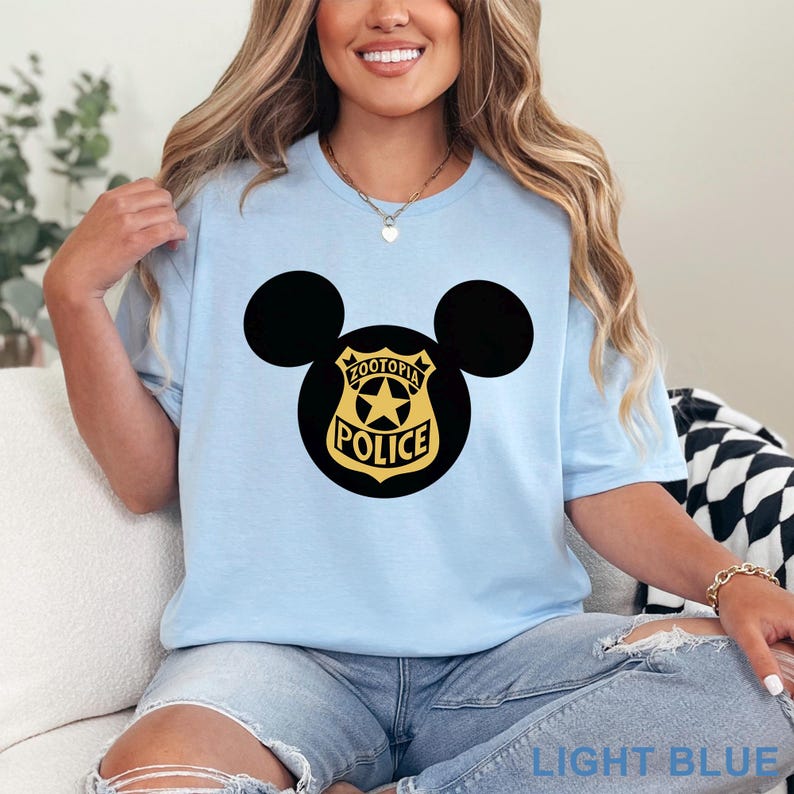 May include: Light blue t-shirt featuring a black Mickey Mouse head design with a Zootopia Police badge in the center. The badge is black with gold lettering and a gold star. The words "LIGHT BLUE" are printed at the bottom of the image.