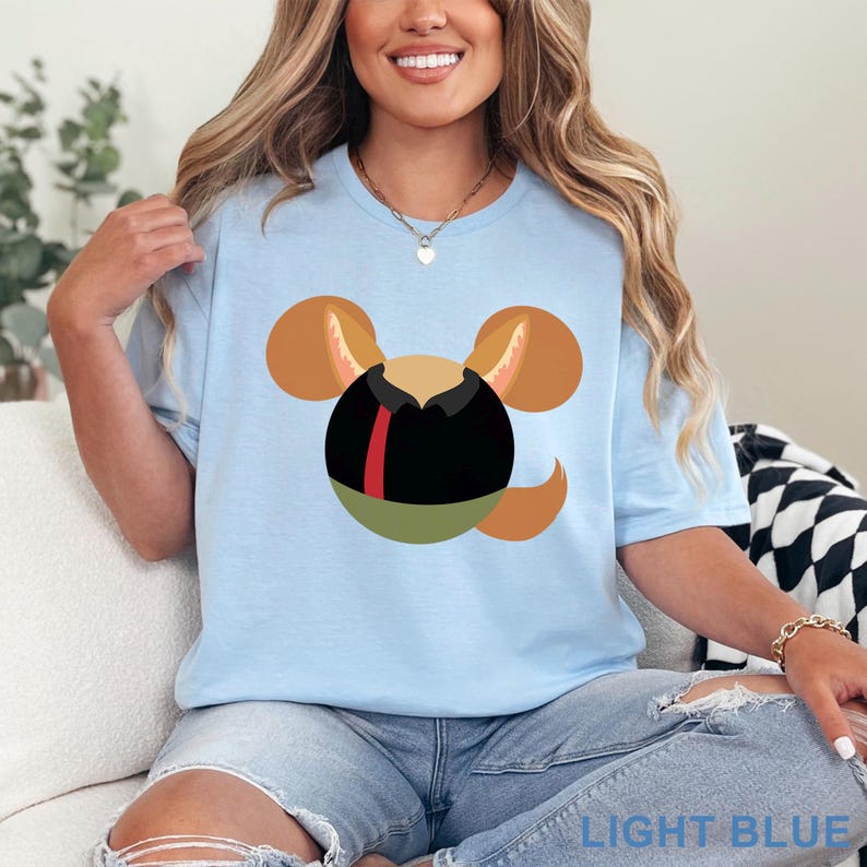 May include: Light blue t-shirt featuring a cartoon character design. The design includes brown ears, a black and green body, and a red stripe. The text "LIGHT BLUE" is printed at the bottom of the shirt. This shirt is a casual wear item.