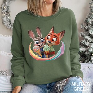 May include: Military green sweatshirt featuring a colorful graphic of Judy Hopps and Nick Wilde from Zootopia holding ice cream cones. The design includes a rainbow arc around the characters. The sweatshirt is a solid color.