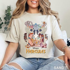 May include: Sand-colored t-shirt featuring a colorful graphic from Disney's Hercules. The design includes Hercules, Megara, and other characters, with the text "Disney's Hercules" in gold lettering. The shirt is made of a soft material.