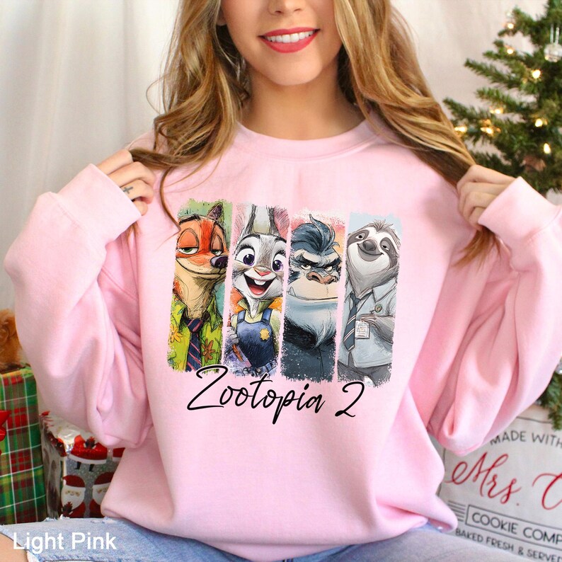 Puede incluir: Sudadera rosa claro con los personajes de Zootopia 2. El dise&ntilde;o incluye a Nick Wilde, Judy Hopps, el jefe Bogo y Flash el perezoso, con el texto "Zootopia 2" en una fuente cursiva.
