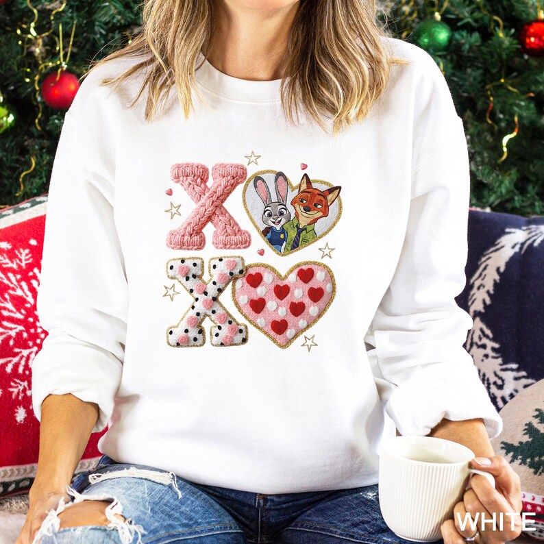 May include: White sweatshirt with embroidered design featuring the characters Judy Hopps and Nick Wilde from Zootopia, along with the letters "X" and "O" and heart shapes. The design includes pink and white colors.