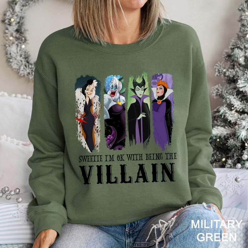 May include: Military green sweatshirt featuring four cartoon villain characters. The text "Sweetie I'm OK with being the Villain" is printed below the characters in black. The sweatshirt is a solid color.
