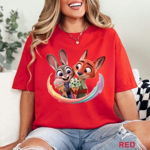 May include: Red t-shirt featuring a colorful graphic of Judy Hopps and Nick Wilde from Zootopia, holding ice cream cones. The design includes a rainbow arc. The word "RED" is printed at the bottom right.