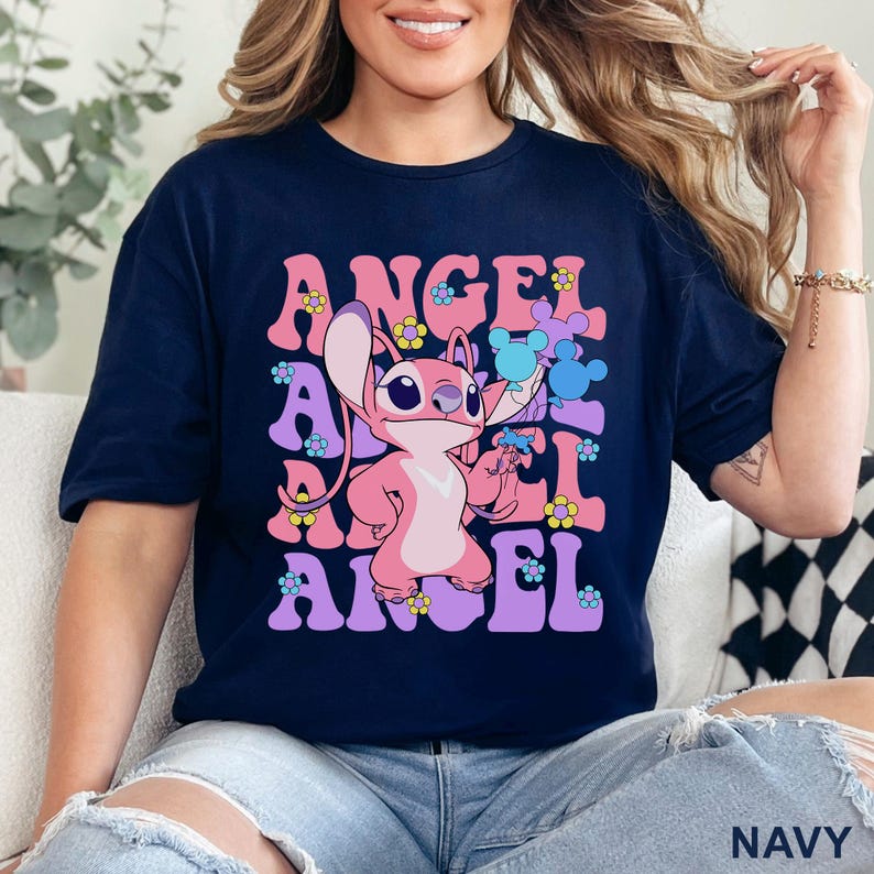 May include: Navy blue t-shirt featuring a pink cartoon character holding puzzle pieces, with the word "ANGEL" in a retro font. The design includes flowers in pink, yellow, and blue. The shirt is a casual wear item.