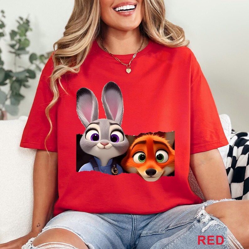 May include: Red t-shirt featuring a graphic of Judy Hopps and Nick Wilde from Zootopia. Judy, a gray bunny, and Nick, an orange fox, are depicted peeking out from a torn-paper effect. The word "RED" is printed in the bottom right corner.