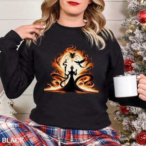 Op de afbeelding: Zwarte sweatshirt met een gestileerde illustratie van een tovenaar met staf, tegen een vurige oranje en gele achtergrond met een volle maan en twee vogels. Het woord "BLACK" staat onderaan gedrukt.