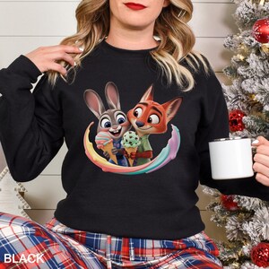 May include: Black crew neck sweatshirt featuring a colorful graphic of Judy Hopps and Nick Wilde from Zootopia holding ice cream cones. The sweatshirt has long sleeves and the word "BLACK" is printed at the bottom.