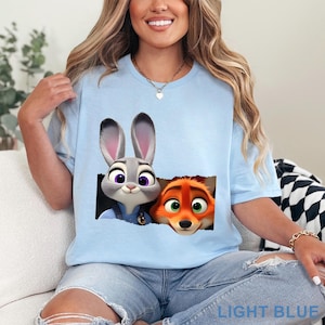 May include: Light blue t-shirt featuring Judy Hopps and Nick Wilde from Zootopia. The graphic shows the characters peeking out of a torn-paper effect. The text "LIGHT BLUE" is printed at the bottom of the shirt.