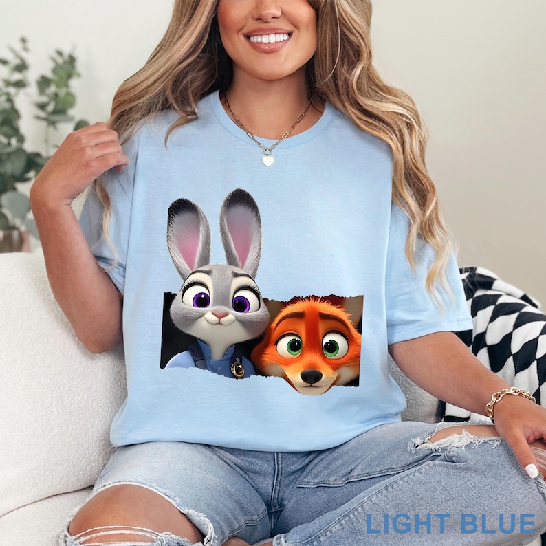 May include: Light blue t-shirt featuring Judy Hopps and Nick Wilde from Zootopia. The graphic shows the characters peeking out of a torn-paper effect. The text "LIGHT BLUE" is printed at the bottom of the shirt.