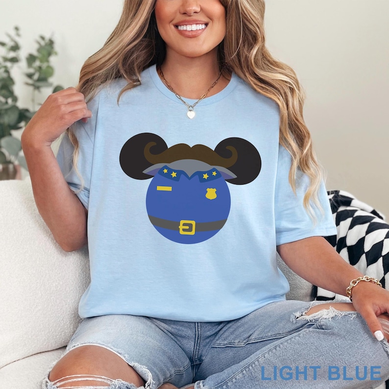 May include: Light blue t-shirt featuring a cartoon character design. The design includes a blue circle with a police officer's hat, mustache, and belt. The text "LIGHT BLUE" is at the bottom of the image.