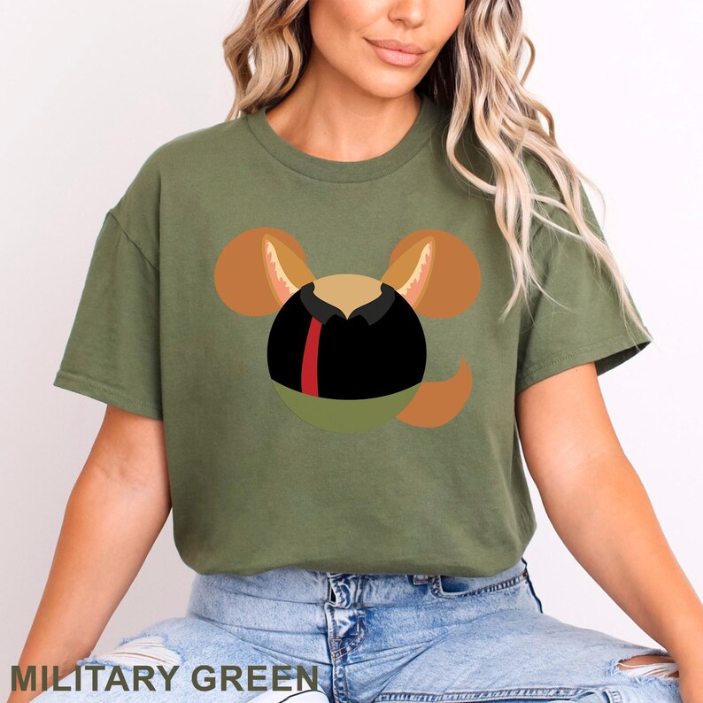 May include: Military green t-shirt featuring a cartoon character design. The design includes a black circle with a red stripe, a green half-circle, and brown ears and tail. The text "MILITARY GREEN" is at the bottom.