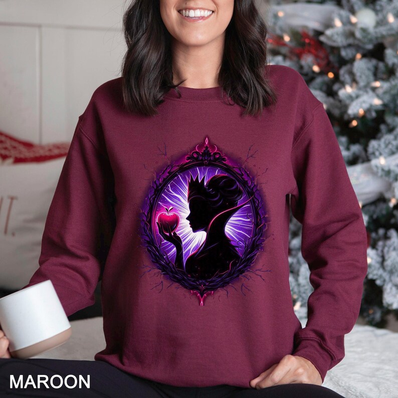 Op de afbeelding: Bordeaux sweatshirt met een silhouet van een koningin die een appel vasthoudt in een decoratief frame. Het ontwerp is in paars en zwart. Het woord "MAROON" staat onderaan het sweatshirt.