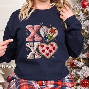 May include: Navy blue sweatshirt with embroidered "XO" design. Features a pink knitted "X", a heart with Judy Hopps and Nick Wilde, and a heart with red polka dots. Gold star accents and the word "NAVY" are also visible.