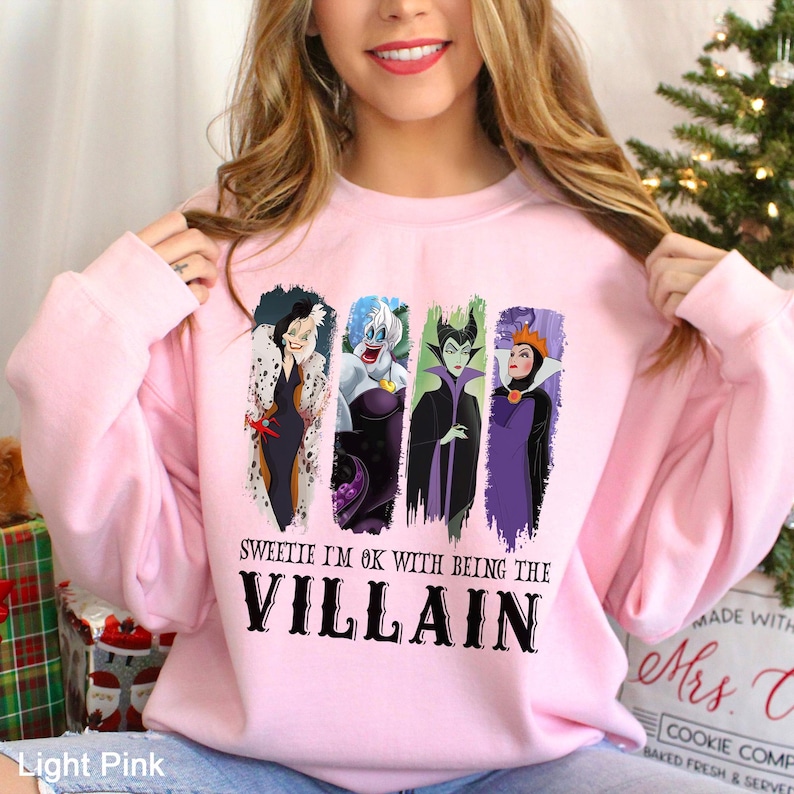 May include: Light pink sweatshirt featuring four cartoon villain characters. The text "SWEETIE I'M OK WITH BEING THE VILLAIN" is printed below the characters in black. The sweatshirt is a light pink color.