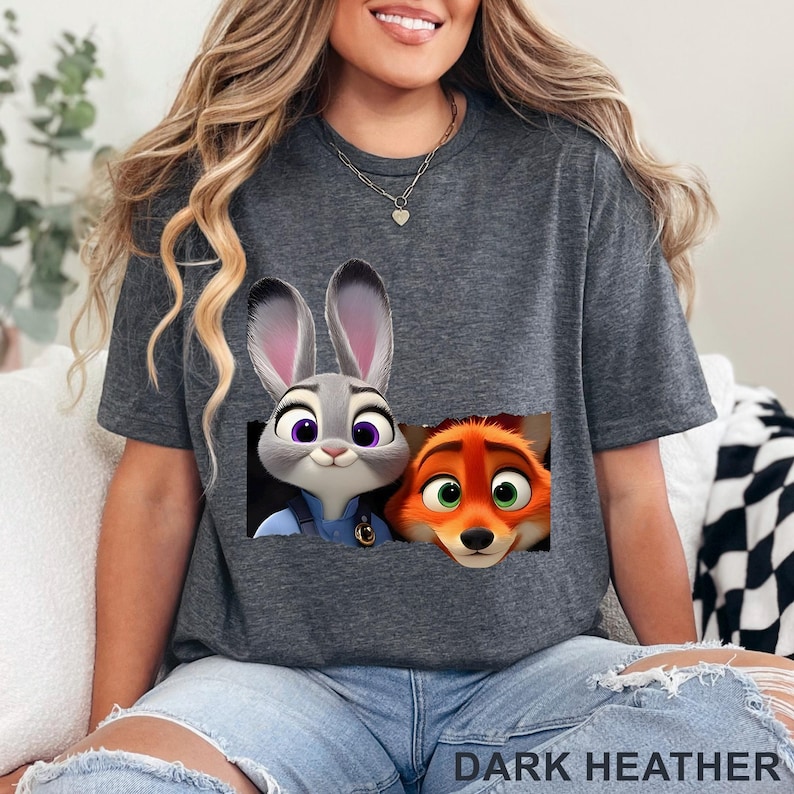 May include: Dark heather gray t-shirt featuring a graphic of Judy Hopps and Nick Wilde from Zootopia. The image shows the characters' faces in a rectangular design. The text "DARK HEATHER" is printed at the bottom of the shirt.