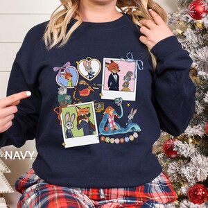 May include: Navy blue sweatshirt featuring multiple cartoon images of characters from Zootopia. The images are arranged in a collage, with characters like Nick Wilde and Judy Hopps in various poses. The word "Zootopia" is also visible.