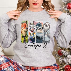 Puede incluir: Una sudadera gris claro con un gr&aacute;fico colorido de personajes de la pel&iacute;cula "Zootopia 2". El dise&ntilde;o incluye a Nick Wilde, Judy Hopps, un gorila y Flash el perezoso. Las palabras "Zootopia 2" est&aacute;n escritas en una fuente cursiva.