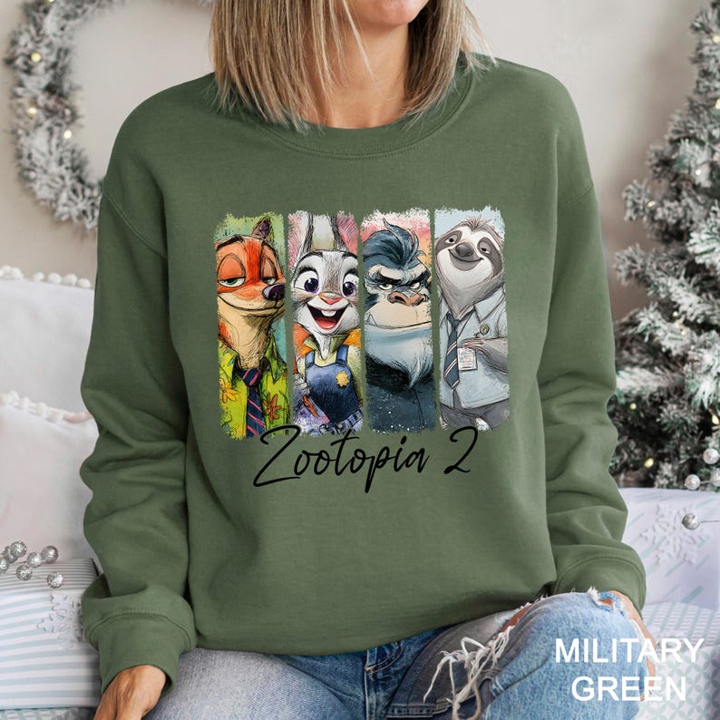 Puede incluir: Sudadera verde militar con un gr&aacute;fico colorido de personajes de Zootopia 2. El gr&aacute;fico incluye a Nick Wilde, Judy Hopps, un gorila y Flash el perezoso. El texto "Zootopia 2" est&aacute; escrito en cursiva debajo de los personajes.