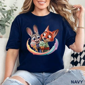 May include: Navy blue t-shirt featuring a colorful graphic of Judy Hopps and Nick Wilde from Zootopia holding ice cream cones. The shirt has a crew neck and short sleeves. The word "NAVY" is printed at the bottom.
