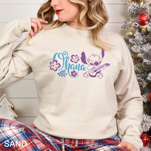 May include: Sand-colored sweatshirt featuring the word "Ohana" in blue script with purple floral accents and a purple cartoon character playing a guitar. The sweatshirt has long sleeves.