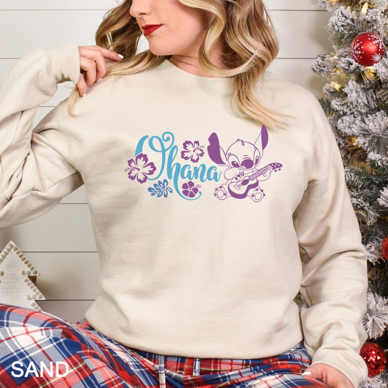 May include: Sand-colored sweatshirt featuring the word "Ohana" in blue script with purple floral accents and a purple cartoon character playing a guitar. The sweatshirt has long sleeves.