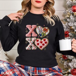 May include: Black sweatshirt with an embroidered design featuring the characters Judy Hopps and Nick Wilde from Zootopia, along with the letters X and O, and hearts. The word "BLACK" is printed at the bottom.