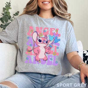 May include: Sport grey t-shirt featuring a pink cartoon character holding blue balloons. The word "ANGEL" is printed in pink and purple, with flower accents. The shirt is made of a soft material.
