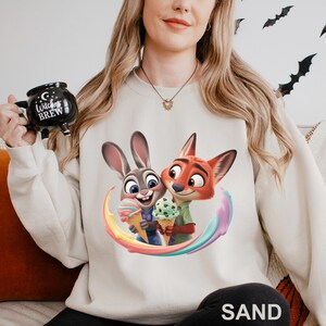 May include: Sand-colored sweatshirt featuring a colorful graphic of Judy Hopps and Nick Wilde from Zootopia holding ice cream cones. The design includes a rainbow arc and the word "SAND" at the bottom. The sweatshirt is long-sleeved.