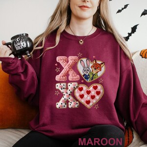 May include: Maroon sweatshirt featuring embroidered "XO" letters and heart designs. One heart contains Judy Hopps and Nick Wilde from Zootopia. The other heart is filled with red hearts. The word "MAROON" is at the bottom.