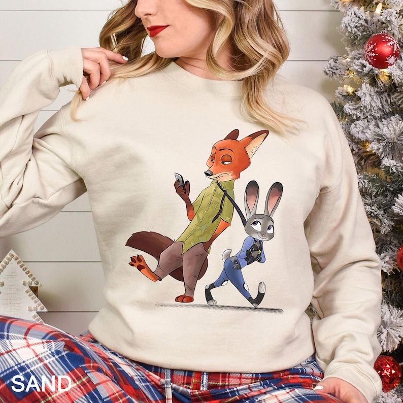 May include: Sand-colored sweatshirt featuring a cartoon illustration of Nick Wilde and Judy Hopps from Zootopia. Nick, a fox, is holding a phone and Judy, a rabbit, is being led by a leash. The word "SAND" is printed at the bottom.