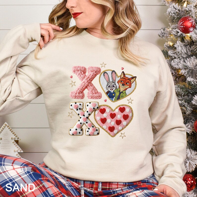 May include: A sand-colored sweatshirt with an embroidered design. The design features the letters "X" and "O", a heart with a cartoon image of a bunny and fox, and a heart with red hearts. The sweatshirt has long sleeves.