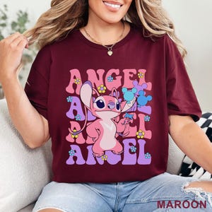 May include: Maroon t-shirt featuring a pink cartoon character holding blue flowers, with the word "ANGEL" in a retro font. The design includes pink, purple, and yellow floral accents. The word "MAROON" is printed at the bottom.