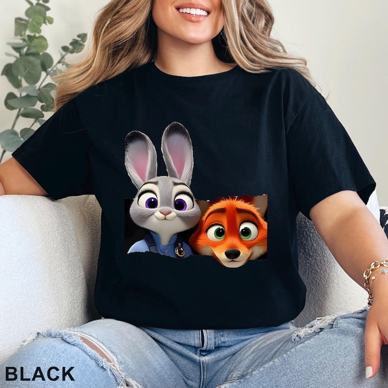 May include: Black t-shirt featuring a graphic of Judy Hopps and Nick Wilde from Zootopia. The cartoon characters are in a pocket-like design. The word "BLACK" is printed at the bottom of the shirt.