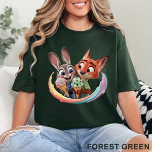 May include: Forest green t-shirt featuring a colorful graphic of the Zootopia characters Judy Hopps and Nick Wilde holding ice cream cones. The image is surrounded by a rainbow arc. The text "FOREST GREEN" is printed at the bottom.